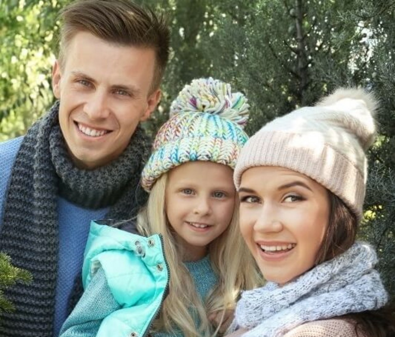 Simbolična slika: mlad par z majhno deklico, v gozdu. Na levi mlad moški, s kratkimi rjavimi lasmi, gleda naprej in se smehlja, v modrem puloverju, okrog vratu temno moder šal. Na sredini: deklica, drobna, v starosti okrog pet let, gleda naprej, na glavi ima pisano kapo z velikim cofom, nosi svetlo turkizno moder brezrokavnik. Na desni: mlada ženska, mama, ki drži deklico, gleda naprej, se smehlja. Na glavi svetla kapa s cofom, okrog vratu svetlo moder šal, oblečena v pulover rožnate barve.