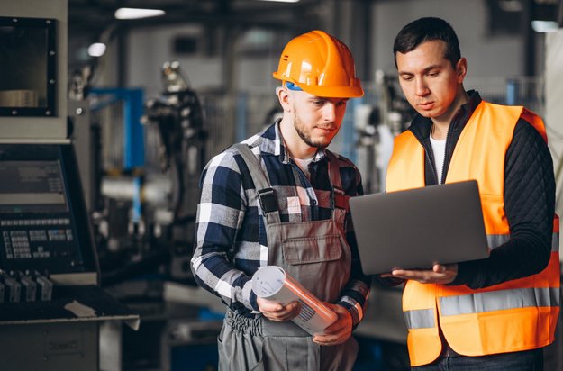 Na desni stojita dva mlajša moška. V ozadju: proizvodno delovno okolje. Moški levo v sivem delovnem kombinezonu in modro-beli srajci, z oranžno čelado na glavi. Zre v temno siv prenosni računalnik, ki ga v rokah drži moški na desni, oblečem v črn pulover in oranžen varnostni jopič.