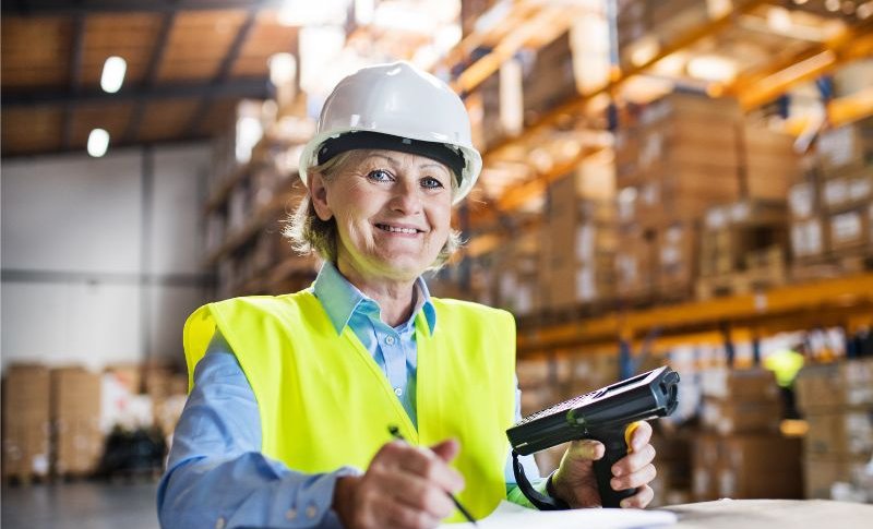 Nasmejana ženska poznih srednjih let s čelado na glavi med delom v skladišču