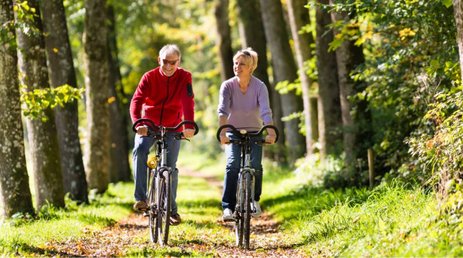 Nasmejan starejši par vštric kolesari po gozdni poti. Na levi moški v rdečem puloverju in kavbojkah, s sončnimi očali. Na desni ženska s kratkimi lasmi, v svetlo vijoličnem puloverju in kavbojkah. 