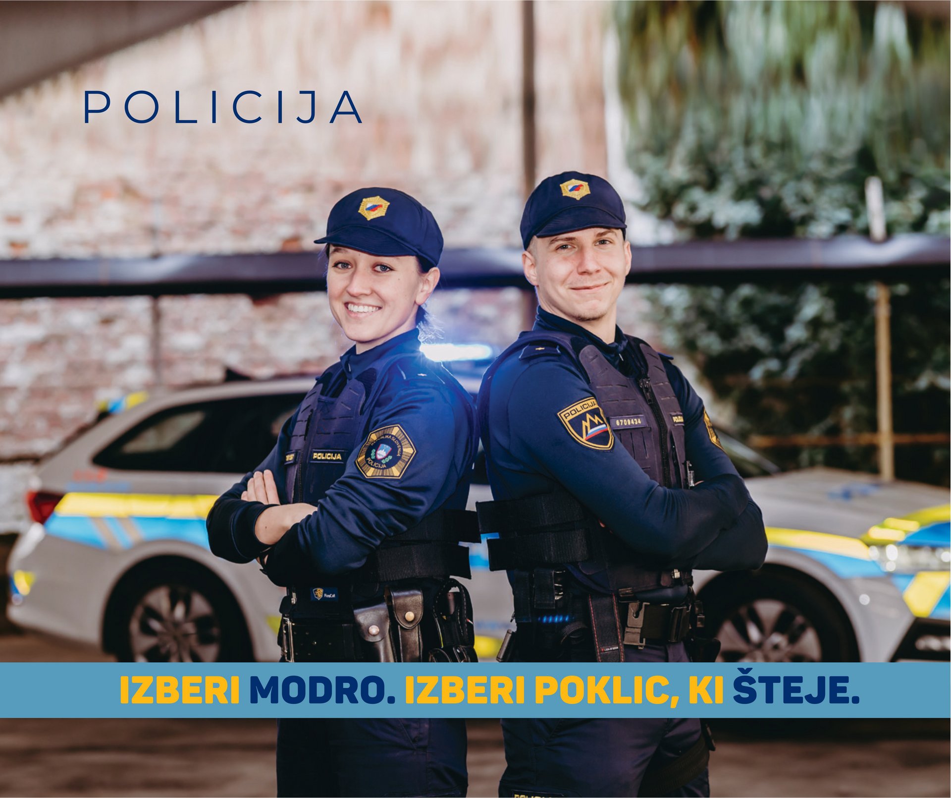 Policist in policistka v uniformi Policije Republike Slovenije, ki stojita pred belim policijskim vozilom.