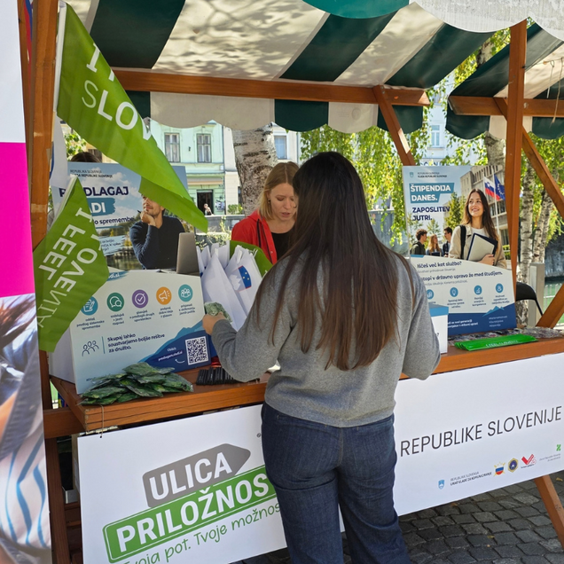 Dogodek Ulica priložnosti - stojnica Urada RS za komuniciranje s promocijskimi izdelki in osebo, ki si ogleduje ponudbo