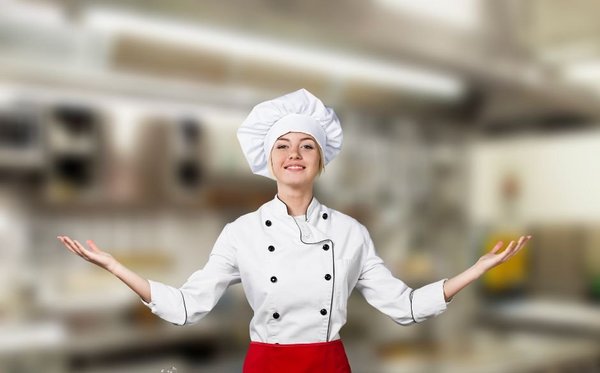 Kuharica: mlajša odrasla ženska, z belo kuharsko kapo na glavi, široko razprtimi rokami in nasmehom na obrazu. Oblečena v belo kuharsko obleko s črnimi gumbi. V ozadju velik kuhinjski obrat. 