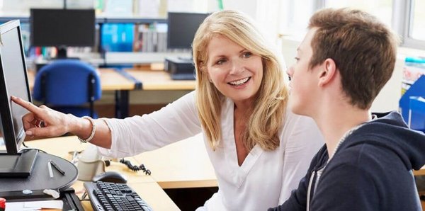 Dekorativna slika: učilnica z računalniki, v ospredju na levi za mizo z računalnikom sedi svetlolasa ženska srednjih let, s prstom kaže na ekran. Zraven nje sedi mlad fant, s kratkimi rjavimi lasmi, gleda v ekran. 