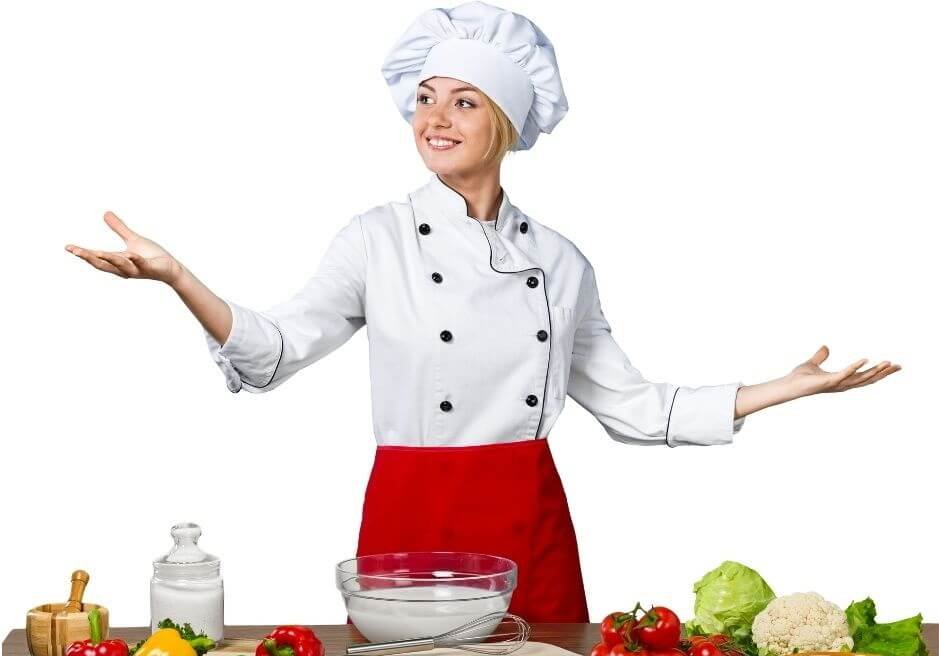 Simbolična slika. Kuharica v beli bluzi in rdečem krilu, z belo kuharsko kapo na glavi, stoji za mizo s sestavinami za pripravo hrane, gleda v levo, z razprtimi rokami, kot da nekoga pozdravlja. 