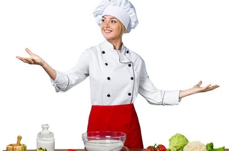 Simbolična slika. Kuharica v beli bluzi in rdečem krilu, z belo kuharsko kapo na glavi, stoji za mizo s sestavinami za pripravo hrane, gleda v levo in se smehlja, s široko razprtimi, dvignjenimi rokami. 
