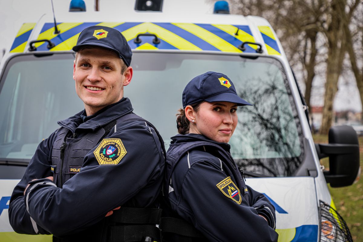 Mlad policist (levo) in mlada policistka (desno), stojita in se stikata s hrbti, v ozadju policijski kombi.