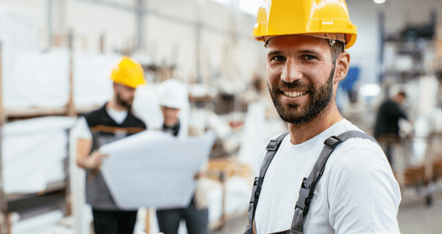 Dekorativna slika za program spodbujanja zaposlovanja v velikih podjetjih: moški v beli majici in modrih delavskih hlačah z naramnicami, z rumeno zaščitno čelado na glavi. V ozadju: delovišče z drugimi delavci.